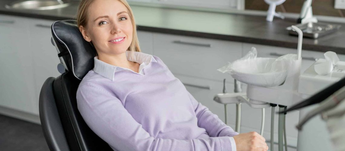 Pretty young smiling female patient of contemporary dentistry sitting in leather armchair and waiting for her doctor in clinics